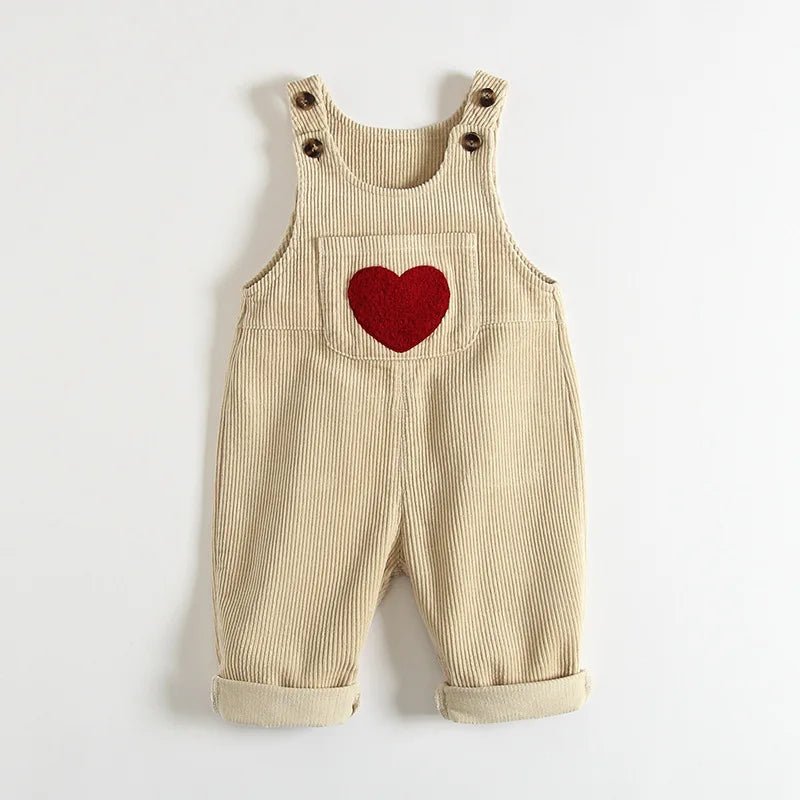 Light beige baby and toddler overalls with a front pocket that has a heart on the front. These baby overall pants have adjustable straps.