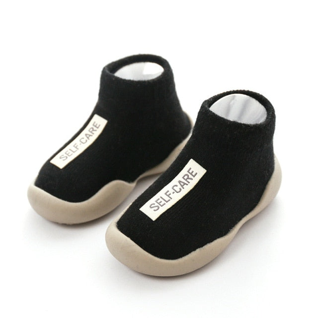 Black first walker sock shoes with a self care lable on the top of the arch.