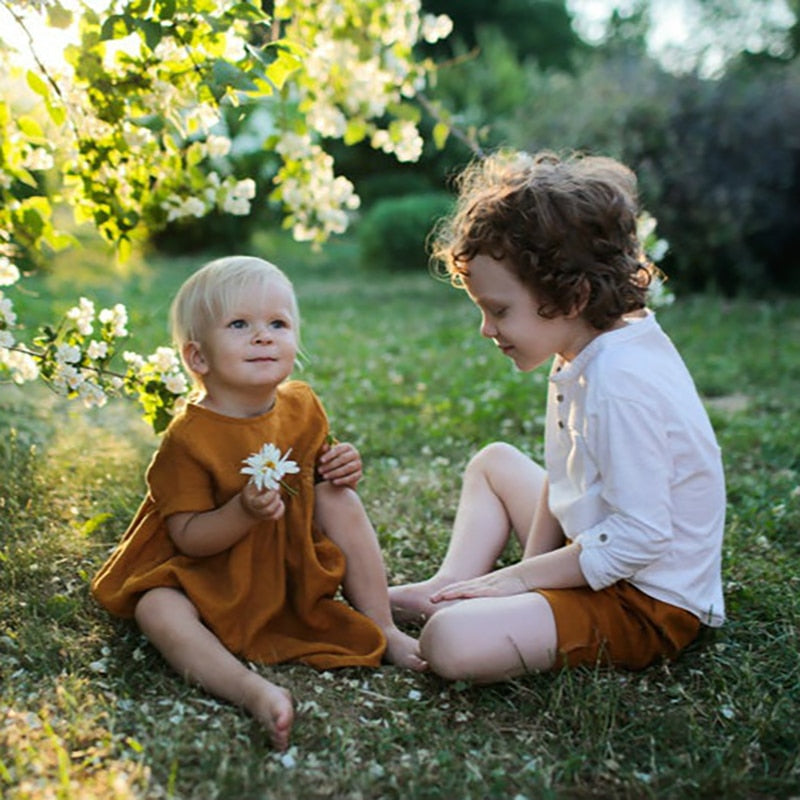 Baby wearing a terracotta summer short sleeved linen dress  and a little boy with curly hair wearing terracotta colored sorts and a white shirt. T>hey are sitting on the grass.